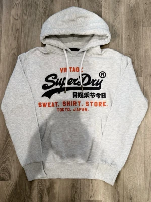 Superdry Vintage Hoodie Grå Strl M - Superdry Vintage grå hoodie med klassisk passform som funkar till allt, storlek M, axelbredd 48 cm, bröstvidd 55 cm, armlängd 62,5 cm, midja 42,5 cm, längd 65 cm, i fint skick.