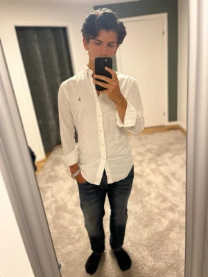 Vit skjorta från Ralph Lauren - Klassisk vit skjorta från Ralph Lauren med knappkrage och broderad logga på bröstet. Skjortan har lång ärm, normal passform och är tillverkad i mjuk bomull. Perfekt till jeans eller chinos för en clean och stilren look.