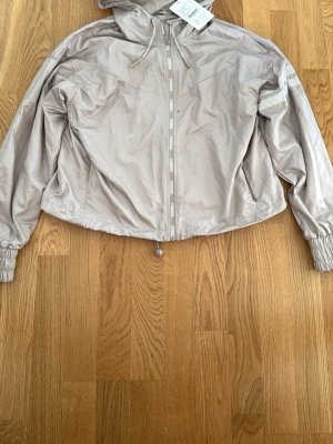 Taupe vindjacka med huva - Snygg taupe vindjacka från Stronger med huva och dragkedja framtill. Jackan har resår i ärmsluten, dragsko i nederkanten och meshfoder på insidan. Detaljer med logga på dragkedja och snören. Perfekt för blåsiga dagar.
