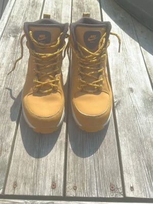 Gula kängor från Nike - Snygga gula kängor från Nike med högt skaft och robust sula. Skorna har gula snören, svarta detaljer runt ankeln och är tillverkade i ett slitstarkt material som liknar nubuck. Perfekta för dig som vill ha en streetig och sportig look.