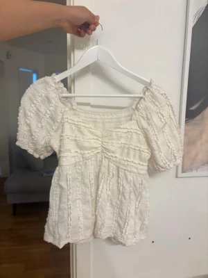Vit blus med puffärm och struktur - Supersöt vit blus med puffiga korta ärmar och strukturerat tyg. Blusen har en lätt rynkad detalj framtill och en luftig, flowy passform. Perfekt för sommarens alla tillfällen och ger en romantisk vibe.