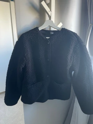 Svart teddyjacka med tryckknappar - Mysig svart teddyjacka med oversized passform och rund hals. Jackan har tryckknappar framtill och är gjord i fluffigt teddy-material som ger en trendig look. Perfekt för dig som vill ha en stilren och enkel jacka till hösten eller vintern. En knapp på fickan har lossnat. 