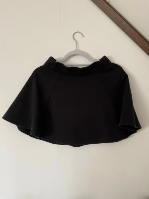 Svart kortkjol med dragkedja - Säljer en svart kortkjol med utställd form och markerad midja. Kjolen har en synlig dragkedja bak och är tillverkad i ett lite tjockare, strukturerat material. Perfekt för dig som gillar en enkel men stilren look.
