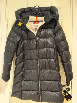 Svart lång Vinterjacka från Parajumpers - Snygg svart lång dunjacka från Parajumpers med stor huva och dragkedja framtill. Jackan har quiltad design, två sidofickor med dragkedja och en patch på ärmen. Perfekt för kalla vinterdagar och riktigt varm tack vare dunfyllningen. Behöver bara en tvätt.