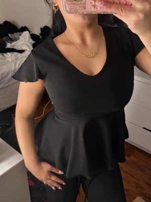 Svart peplum topp med v-ringning - Snygg svart peplum topp med v-ringning och korta ärmar. Toppen har en markerad midja och ett utställd volang nedtill som ger en feminin siluett. Perfekt att styla med jeans eller kjol för en trendig look.