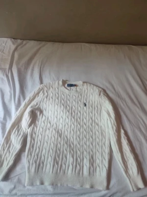 Vit kabelstickad tröja Polo Ralph Lauren - Kabelstickad vit tröja från Polo Ralph Lauren med rund halsringning och broderad logga på bröstet. Tröjan har långa ärmar och ribbade muddar vid ärmslut och nederkant. Perfekt för en klassisk och stilren look.
