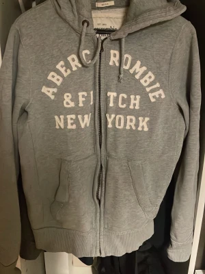 Grå hoodie Abercrombie & Fitch - Grå zip-hoodie från Abercrombie & Fitch med vit broderad text 'New York' på bröstet. Klassisk modell med huva, dragsko och två fickor framtill. Perfekt för en avslappnad och cool stil. Storlek XL men den sitter snarare som en M med loose fit. Skriv om ni har frågor😊