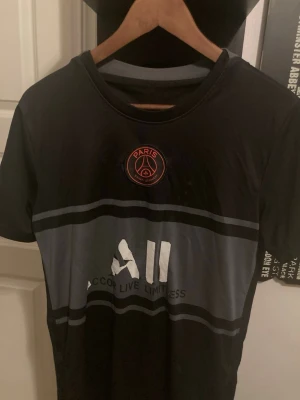 Svart PSG t-shirt med tryck - Säljer en svart PSG t-shirt med klubbens emblem i rött på bröstet och texten 'Accor Live Limitless' framtill. T-shirten har rund hals, korta ärmar och gråa detaljer över bröstet. Perfekt för dig som gillar fotboll och vill visa ditt stöd för Paris Saint-Germain.