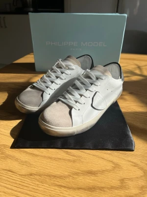 Vita Philippe Model sneakers - Snygga vita sneakers från Philippe Model med beige mockadetaljer på tån och klassisk logotyp på sidan. Skorna har vita skosnören, låg profil och en stilren design i läder och mocka. Perfekta för dig som gillar minimalistisk och trendig stil.