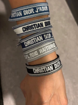 Christian Dior J'adior tygarmband - Säljer flera snygga tygarmband från Christian Dior med texten 'J'adior'. Finns i olika färger som blå, grå, svart och vit. Armbanden har en cool, vävd design och passar perfekt för dig som vill ha en trendig accessoar med lyxig känsla.