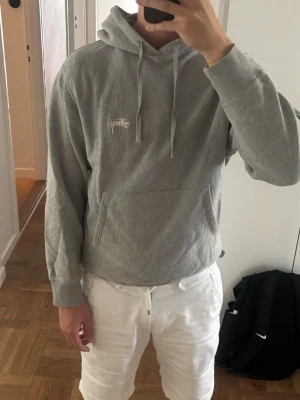 Grå hoodie från Stussy - Säljer en stilren grå hoodie från Stussy med klassisk logga broderad på bröstet. Hoodien har huva med dragsnören, stor magficka och ribbade muddar vid ärmar och midja. Perfekt för en avslappnad streetwear-look.