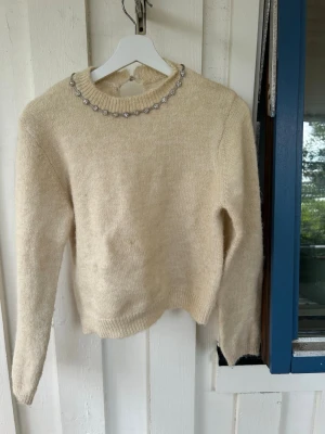 Beige stickad tröja med pärldetaljer - Mysig beige stickad tröja med långa ärmar och ribbade muddar. Rund halsringning dekorerad med pärlliknande blommor. Baksidan har en liten öppning med knappstängning vid nacken. Perfekt för dig som gillar en söt och stilren look.
