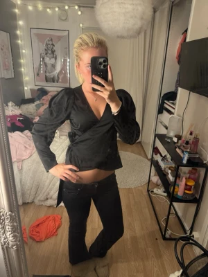 Svart omlottblus med puffärm - Snygg svart omlottblus i glansigt material med puffiga axlar och långa ärmar. Knytband i midjan för en figurnära passform och djup v-ringning framtill. Perfekt att styla med jeans eller kjol för en trendig look.
