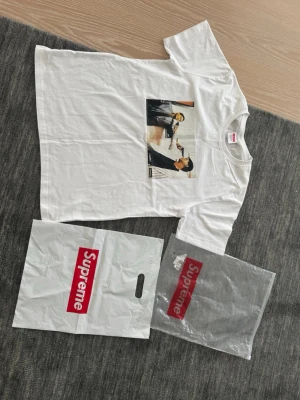 Vit Supreme The Killer T-shirt - Supreme T-shirt i vitt med tryck från filmen The Killer på framsidan och citat på ryggen. Klassisk box logo-påse medföljer. T-shirten har rund hals och korta ärmar, tillverkad i mjuk bomull. Perfekt för dig som gillar streetwear och ikoniska prints.