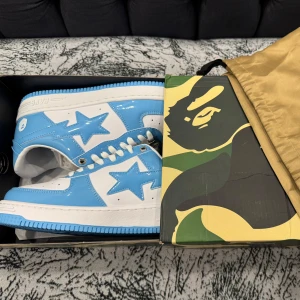BAPE STA sneakers blå och vita - Säljer ett par BAPE STA sneakers i en fräsch blå och vit färgkombination. Skorna har en glansig finish, klassisk stjärndetalj på sidan och vit snörning. Kommer i originalkartong med camo-mönster. Perfekt för dig som vill sticka ut med streetwear.