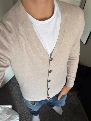 Beige tunn cardigan med knappar - Stilren beige cardigan med v-ringning och svarta knappar framtill. Tillverkad i ett tunt, mjukt material som passar perfekt över en t-shirt. Långärmad modell med ribbade muddar och nederkant. Enkel att matcha till olika outfits.