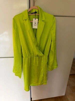 Limegrön omlottklänning från BikBok - Säljer en limegrön kort omlottklänning från BikBok med lång ärm och krage. Klänningen har en snygg omlottdesign och är tillverkad i ett glansigt material som ger en cool look. 