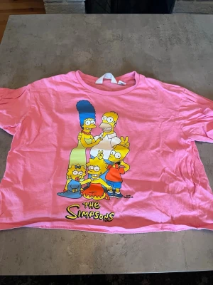 Rosa The Simpsons t-shirt från H&M - Säljer en rosa t-shirt från H&M med stort tryck av The Simpsons-familjen på framsidan. T-shirten har korta ärmar, rund hals och är gjord i mjuk bomull. Perfekt för dig som älskar tecknade serier och vill ha en färgglad look.