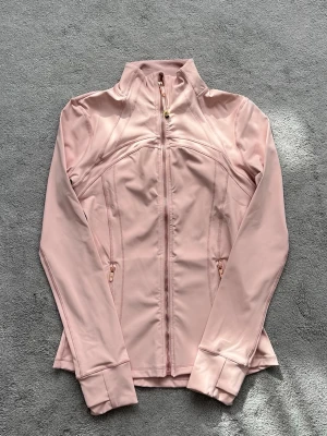 Ljusrosa träningsjacka från Lululemon - Snygg ljusrosa träningsjacka från Lululemon med hel dragkedja framtill, hög krage och två fickor med dragkedja. Tillverkad i ett stretchigt och mjukt funktionsmaterial som passar perfekt till träning eller som ett extra lager. Tumhål vid ärmsluten för extra komfort.