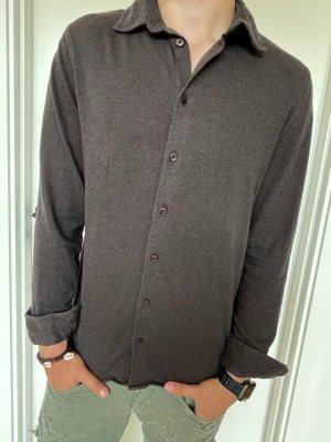 Mörkbrun långärmad skjorta - Stilren mörkbrun skjorta med klassisk krage och knappar framtill. Skjortan har lång ärm och en rak passform som ger en avslappnad look. Tillverkad i mjukt bomullsmaterial som känns skönt mot huden. Perfekt för dig som gillar enkel och clean stil. Nypris 799kr modellen är 179cm lång