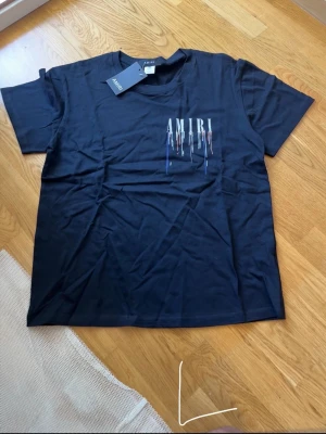 Svart AMIRI t-shirt med tryck - Snygg svart t-shirt från AMIRI med färgstarkt tryck framtill och baktill där loggan droppar i blått, vitt och rött. Klassisk passform och rund hals. Perfekt för dig som vill ha en statement tee med cool design.