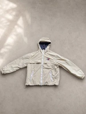 Beige vindjacka från Tommy hilfiger - Snygg beige vindjacka från Tommy Jeans med huva och vita dragkedjor. Jackan har meshfoder, två fickor med dragkedja och logga på bröstet. Perfekt för blåsiga dagar och enkel att matcha med olika outfits. Använd fåtal gånger.