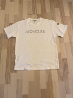 Vit Moncler t-shirt med logga - Säljer en klassisk vit t-shirt från Moncler med stor logga tryckt över bröstet och en liten patch på ärmen. T-shirten har rund hals och korta ärmar. Perfekt till jeans eller shorts för en clean och stilren look.