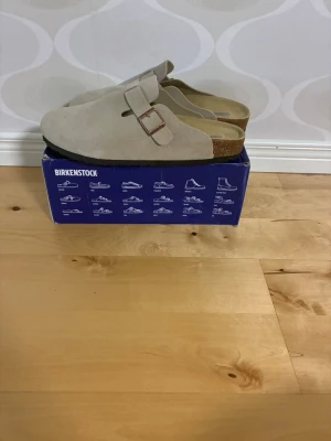Beige Birkenstock tofflor mocka - Säljer ett par beigea Birkenstock tofflor i mocka med klassisk slip-in design och justerbart spänne i metall. Fotbädden är ergonomisk och innersulan är i mjukt material för extra komfort. Perfekta för chill dagar hemma eller ute.