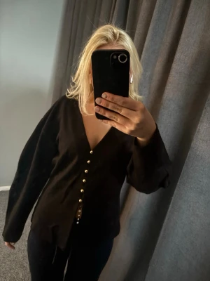 Svart blus med guldknappar - Stilren svart blus med djup v-ringning och dekorativa guldknappar framtill. Blusen har långa ärmar och en något lös passform, perfekt för en trendig look. Materialet känns mjukt och lätt mot huden.