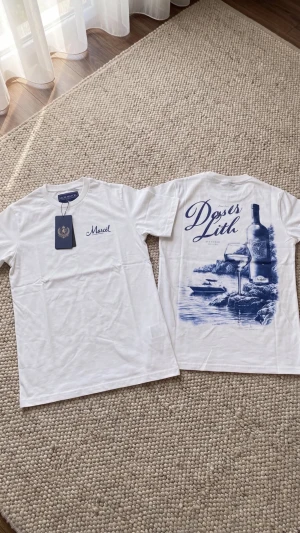 Vit t-shirt med blått tryck - Vit t-shirt med blått tryck fram och bak. På bröstet står det 'Marcel' och på ryggen finns ett stort motiv med texten 'Doss Lith' samt illustration av vinflaska, glas och havsmotiv. Klassisk passform och rund hals.