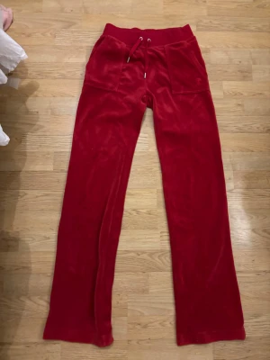 Röda Juicy Couture byxor - Mysiga röda mjukisbyxor i velour med raka ben, bred resår i midjan och dragsko. Stora fickor framtill och en broderad bokstavsdetalj på ena fickan.  Använda några få tillfällen