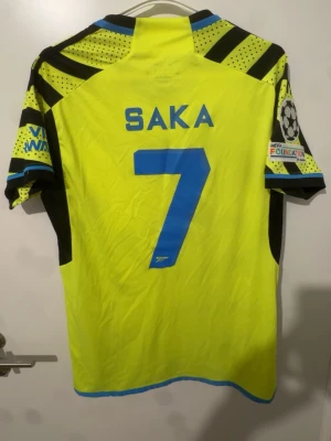 Arsenal Saka matchtröja gul/svart - Cool Arsenal matchtröja i gult och svart med blå detaljer, Saka 7 på ryggen. Tröjan har tryckta loggor, Champions League-märke på ärmen och sponsortryck framtill. Tillverkad i lätt och ventilerande material, perfekt för fotboll eller träning.