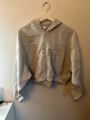 Grå croppad hoodie med dragkedja - Säljer en ljusgrå croppad hoodie med luva och dragkedja framtill. Hoodien har resår i midjan och vid ärmsluten för en skön och avslappnad passform. Perfekt för dig som gillar en enkel och clean stil.