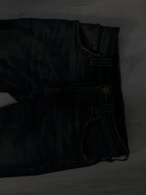 Nudie jeans kan få mer bilder på dem - Säljer ett par mörkblå slim fit jeans med klassisk femficksdesign och snygga slitningar. Jeansen har guldfärgade sömmar och metallknapp i midjan. Perfekta för dig som gillar en smalare passform och tidlös look.