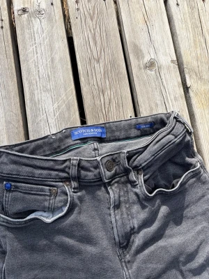 jeans från Scotch & Soda - Snygga gråsvarta jeans från Scotch & Soda med klassisk femficksdesign och knappgylf. Jeansen har en tvättad look och smal passform som sitter tajt längs benen. Perfekta för en trendig och avslappnad stil. Mycket feta detaljer och snygg tvätt.