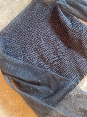 Svart mesh topp med puffärmar - Trendig svart topp med smockad kropp och transparenta meshärmar. Puffiga långärmar i tunn mesh med diskreta prickar ger en cool och modern look. Perfekt att styla med jeans eller kjol för en edgy vibe.