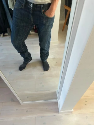 Mörkblå jeans - Säljer ett par mörkblå jeans med rak passform och klassisk femficksdesign. Jeansen har kontrastsömmar och normal midja. Materialet är denim i bomull och de har en snygg tvättad look. Perfekta för dig som gillar en enkel och stilren stil.