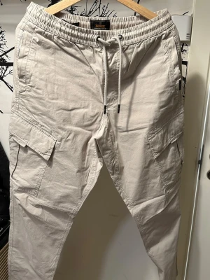 Beige cargobyxor med dragsko - Oanvända, Snygga beige cargobyxor med avslappnad passform och dragsko i midjan. Byxorna har stora fickor på sidorna och är tillverkade i ett mjukt bomullsmaterial. Perfekta för en casual streetwear-look.