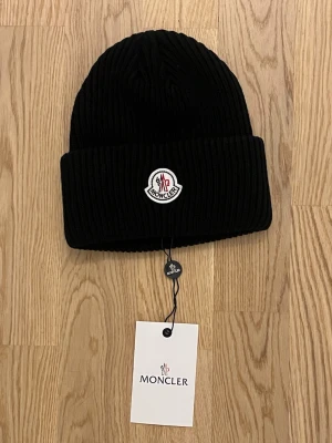 Svart ribbstickad mössa Moncler - Snygg svart ribbstickad mössa från Moncler med uppvikt kant och klassisk Moncler-logga framtill. Tillverkad i mjukt material som håller värmen och ger en clean look. Perfekt accessoar för kalla dagar och enkel att matcha med olika outfits.