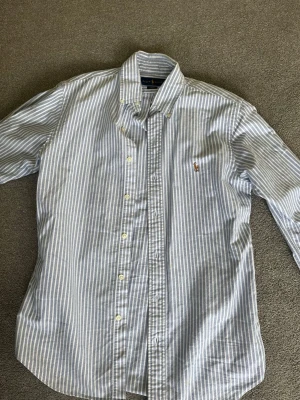 Randig skjorta från Ralph Lauren - Klassisk blå- och vitrandig skjorta från Ralph Lauren i slim fit-modell. Skjortan har långa ärmar, knappar framtill och den ikoniska broderade loggan på bröstet. Tillverkad i mjuk bomull för en skön känsla.