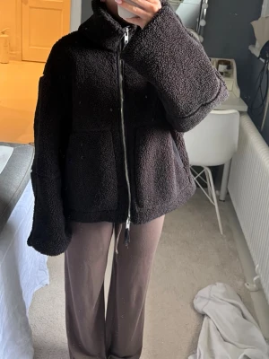 Svart oversized teddyjacka med dragkedja - Mysig svart teddyjacka med oversized passform och hög krage. Jackan har två stora fickor framtill och stängs med en silverfärgad dragkedja. Perfekt för kalla dagar när du vill vara både varm och stilren.
