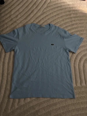 Ljusblå t-shirt från Lacoste - Säljer en stilren ljusblå t-shirt från Lacoste med den klassiska krokodilloggan broderad på bröstet. T-shirten har rund halsringning och korta ärmar. Perfekt basplagg för en clean och fräsch look.