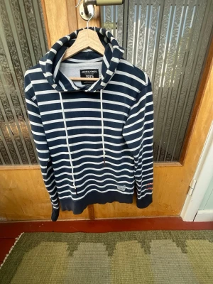 Randig hoodie från Jack & Jones - Mörkblå hoodie med vita horisontella ränder från Jack & Jones. Tröjan har stor huva med långa vita snören och ribbade muddar vid ärmslut och nederkant. Perfekt för en avslappnad och stilren look.
