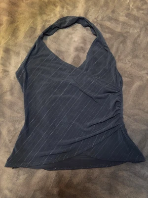 Svart omlott halterneck topp - Snygg svart  halterneck topp med omlott-design och diskret randigt mönster. Toppen har v-ringning och rynkade detaljer på sidan för en cool look. Perfekt för dig som vill ha något stilrent men ändå unikt.