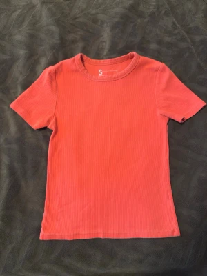 Ribbad korallrosa t-shirt - En enkel och stilren ribbad t-shirt i en snygg korallrosa färg. T-shirten har korta ärmar, rund halsringning och är gjord i ett mjukt bomullsmaterial. Perfekt till jeans eller kjol för en avslappnad look.