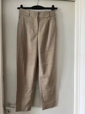 Beige linnebyxor med hög midja - Snygga beige linnebyxor med hög midja och raka ben. Byxorna har breda hällor för bälte, pressveck framtill och två dekorativa bakfickor. Perfekta för en stilren och avslappnad look.