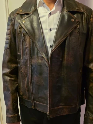 Brun biker skinnjacka med dragkedjor - Snygg mörkbrun skinnjacka i bikerstil med flera dragkedjor framtill och detaljer på axlarna. Jackan har bred krage med tryckknappar och en cool, sliten finish som ger en vintagekänsla. Perfekt för dig som vill ha en edgy look.