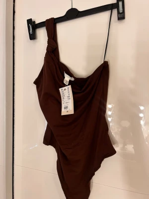 Brun oneshoulder stringbody med knut - Stilren brun linnetopp med oneshoulder och snygg knutdetalj på axeln. Toppen är tight och ärmlös, tillverkad i mjuk bomull som sitter skönt mot kroppen. Helt oanvänd med prislapp 