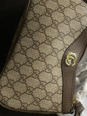 Beige monogram handväska med guldspänne - Snygg beige handväska med klassiskt monogrammönster i brunt och detaljer i brunt läder. Väskan har ett elegant guldspänne och axelrem. Perfekt för dig som gillar tidlös och trendig design.