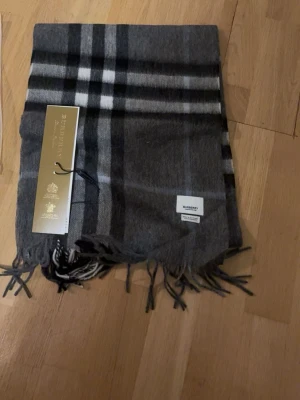 Rutig grå halsduk - Snygg grå halsduk  med klassiskt rutmönster i svart, vitt och grått. Halsduken har fransar i ändarna och är tillverkad i mjukt material. Perfekt accessoar för att lyfta din outfit speciellt när vintern närmar sig 🙃